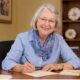 Elizabeth H. Cottrell, silver-haired and wearing a blue shirt and floral scarf, smiling as she writes a note by hand at a wooden desk with envelopes and flowers.